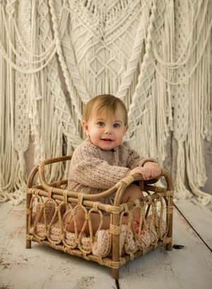 Smiling baby in knit outfit sitting in rattan crib with macrame backdrop on rustic white wood floor.