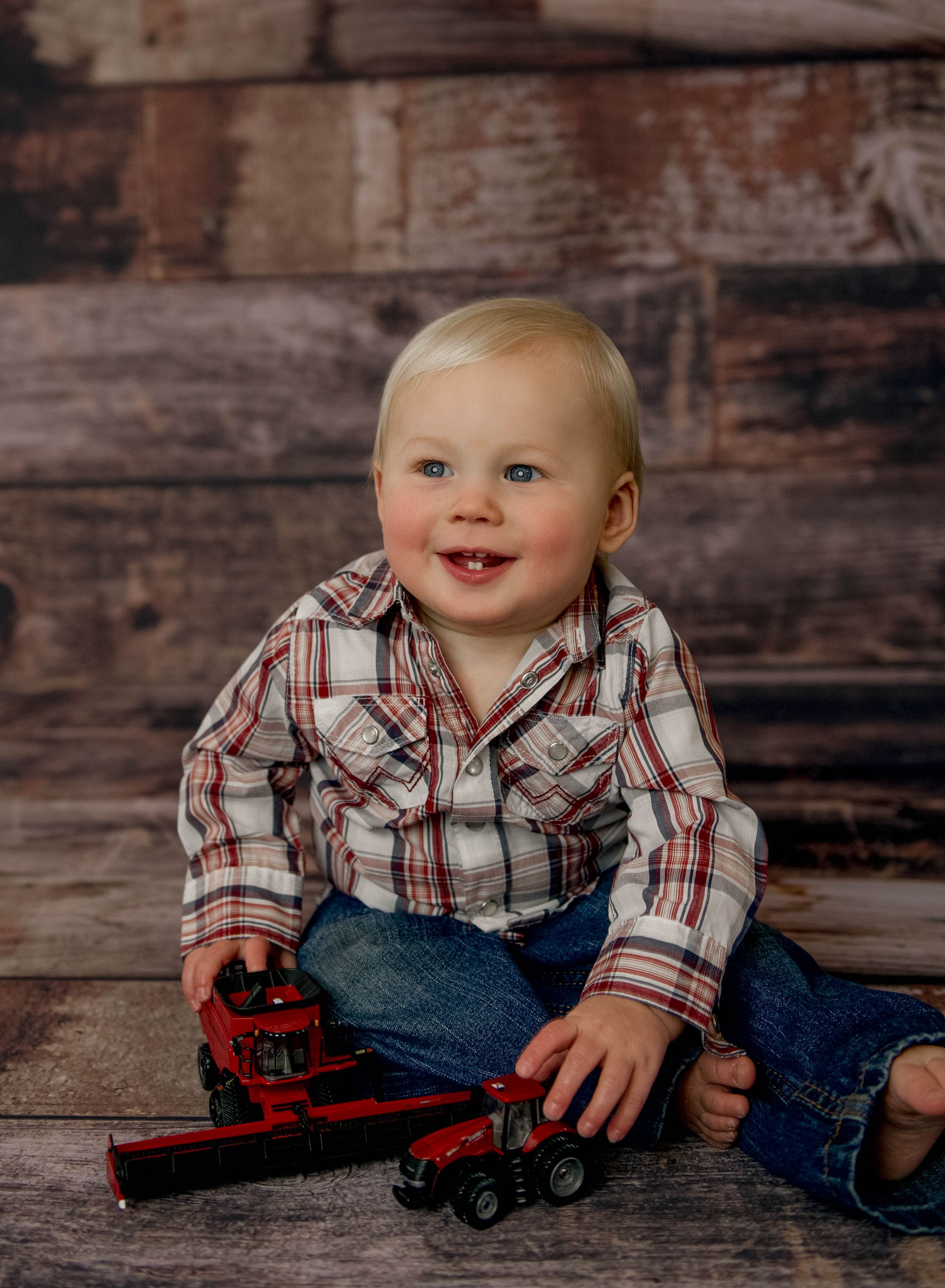 Smiling toddler boy in plaid shirt and jeans sitting on floor with red toy farm equipment against rustic wood backdrop.