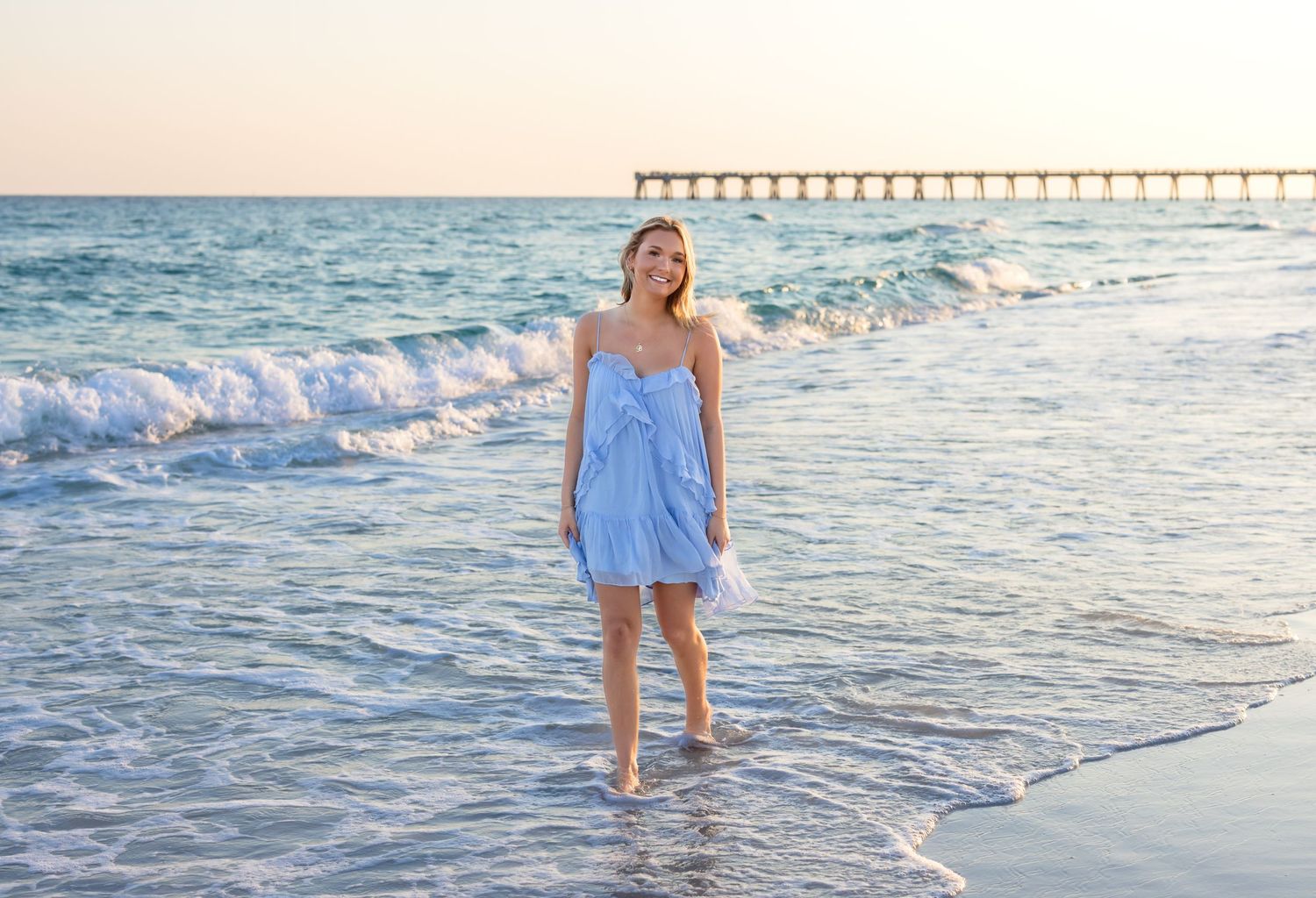 Woman in blue sundress standing barefoot on sandy beach with ocean waves and pier in background.