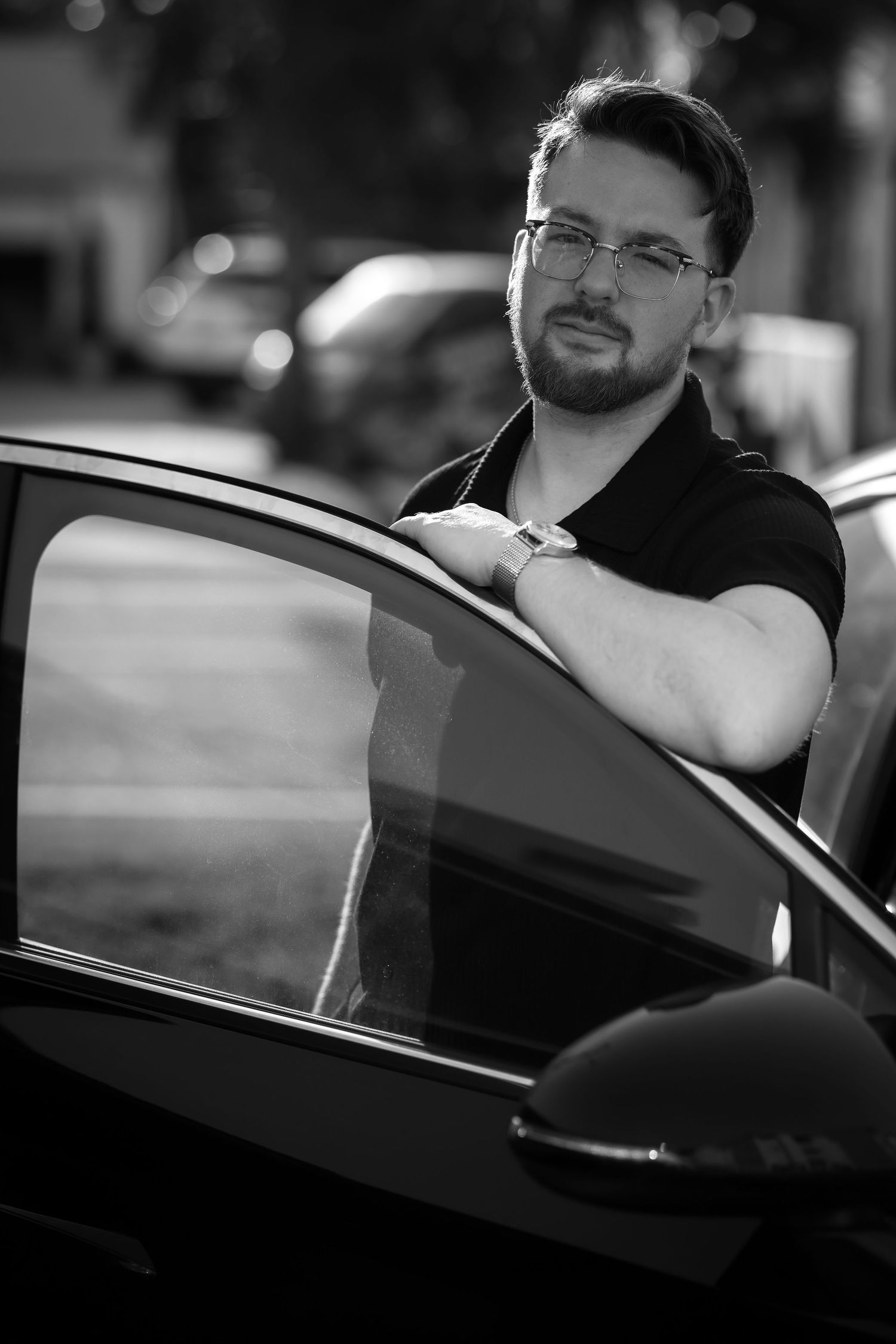 Black and white portrait of a person wearing glasses leaning against an open car door in natural lighting.