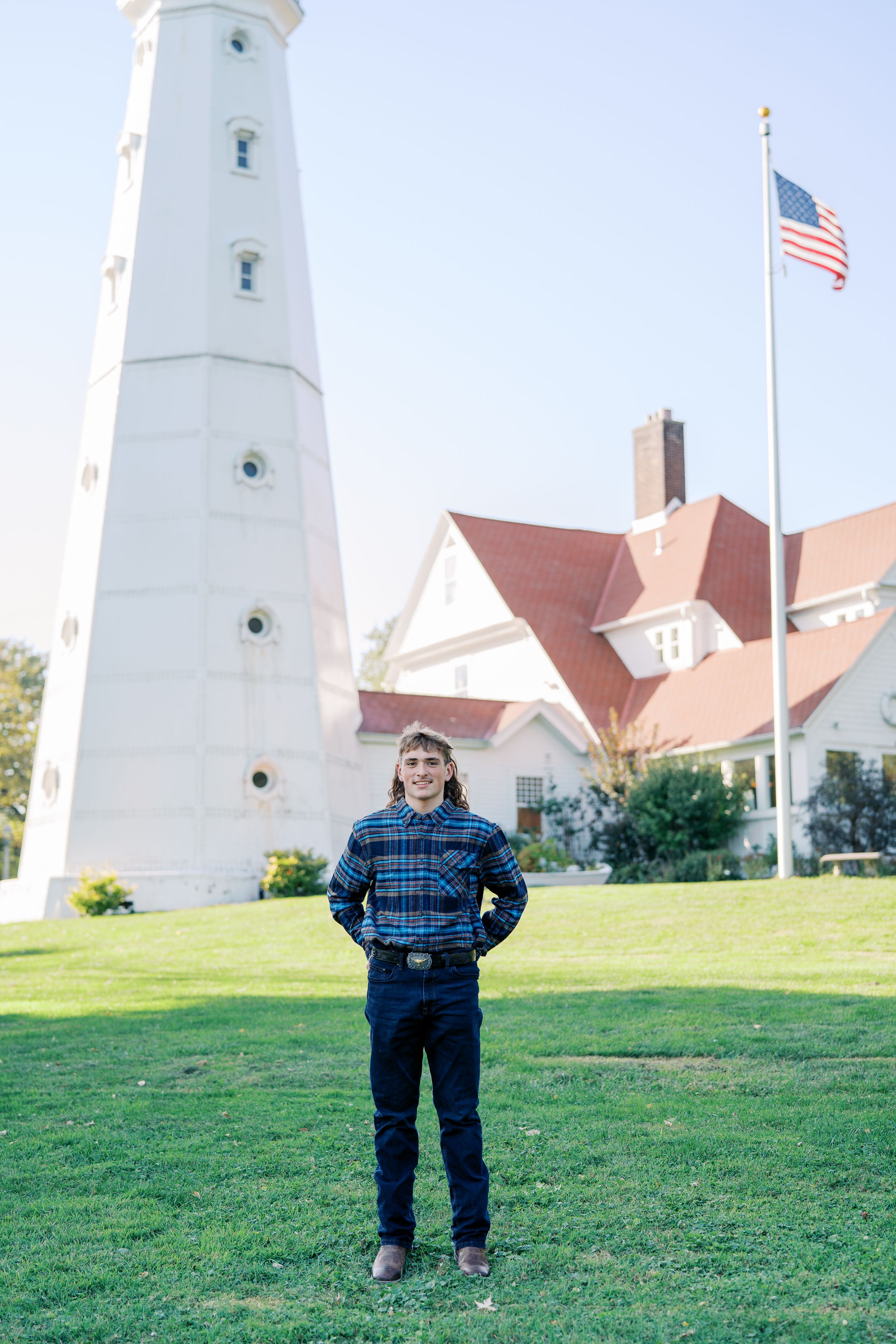 Person in plaid shirt stands in front of white lighthouse and American flag.
