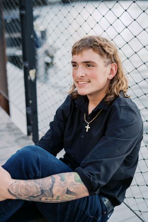 Casual poses against chain link fence wearing dark clothing and cross necklace.