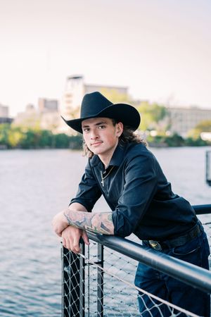 Person in dark clothing and cowboy hat leans on waterfront railing with cityscape behind.