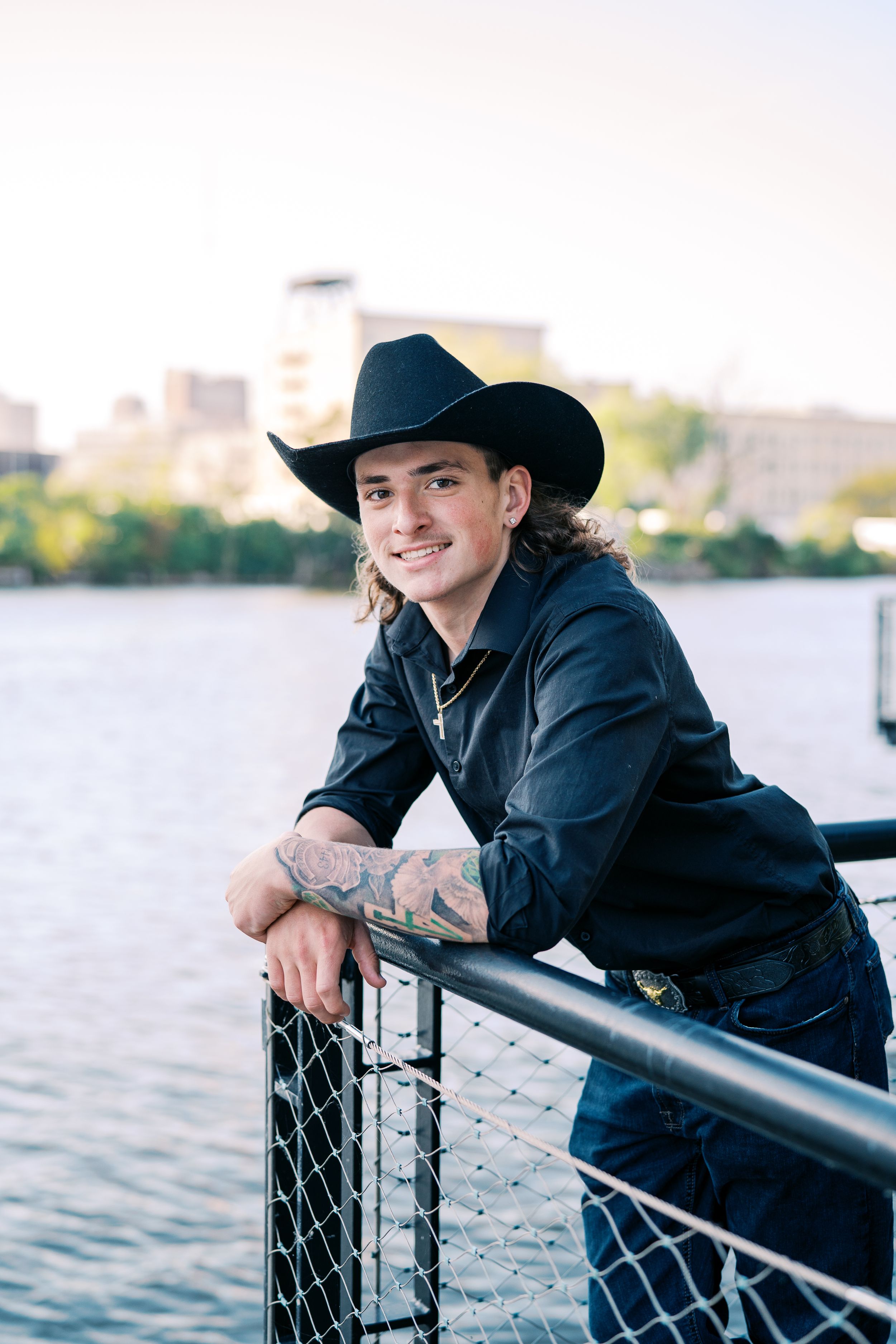 Cowboy in black attire leans against railing by waterfront with cityscape in background.