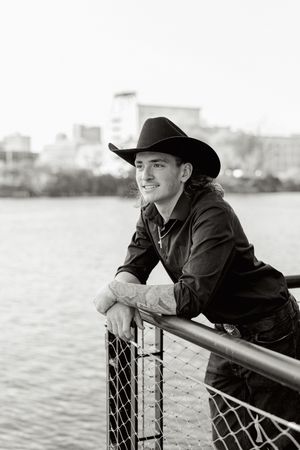 Black and white portrait of cowboy leaning on railing near water.