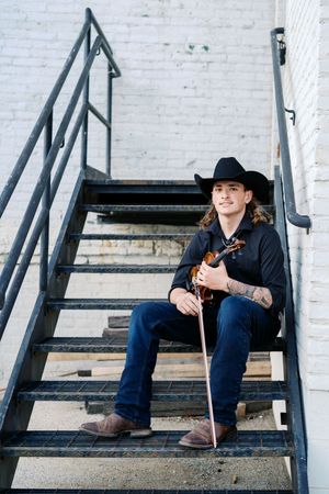 Musician in western wear sits on metal staircase with guitar.