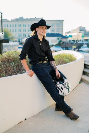 Western-styled figure in dark clothing poses against urban backdrop.