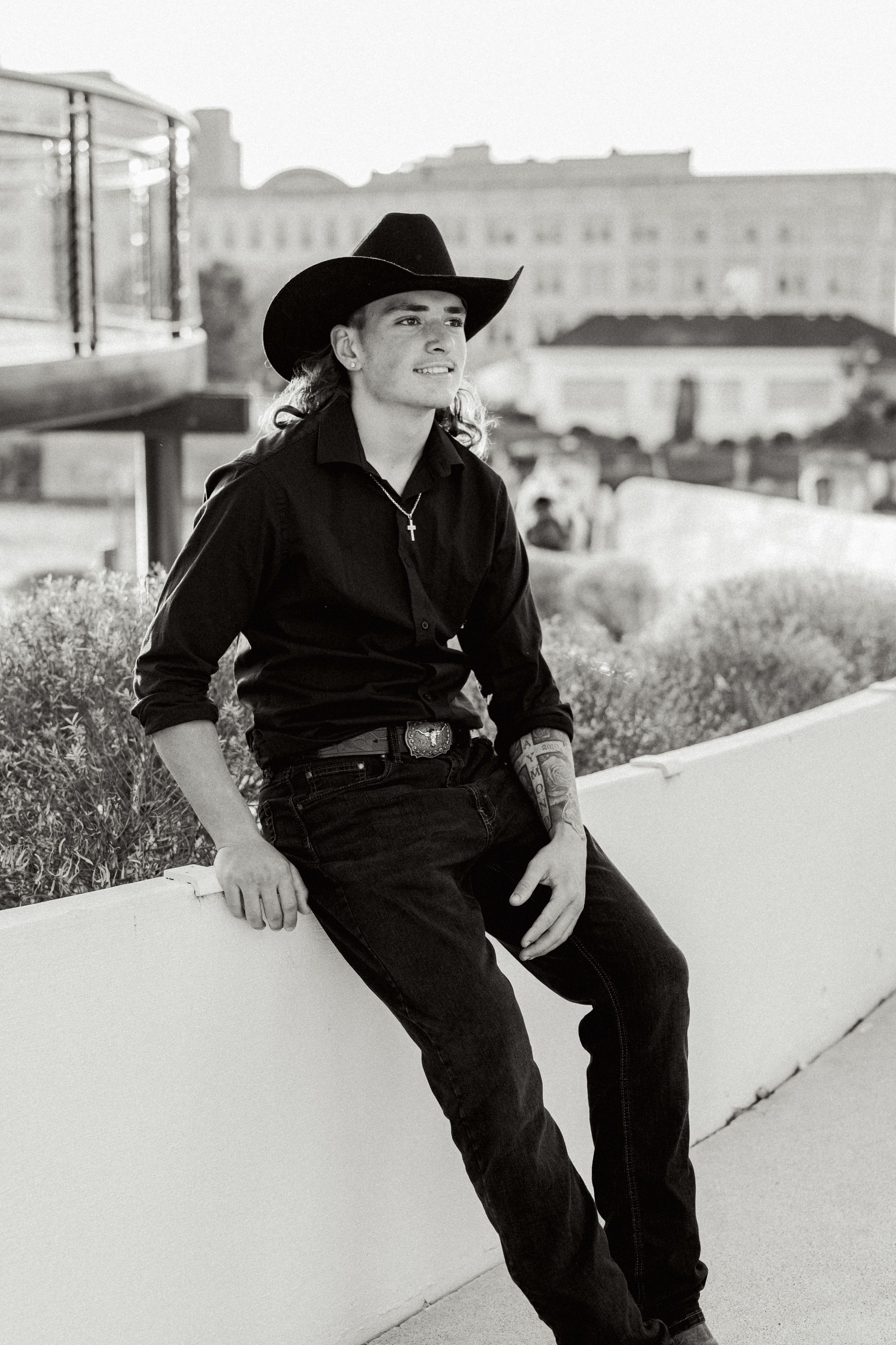 Black and white portrait of cowboy sitting on concrete wall with city buildings behind.