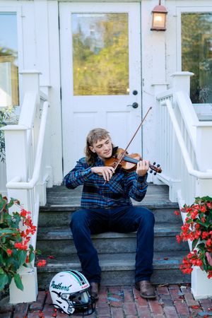 Person in plaid shirt plays violin on white porch with flowers and football helmet.