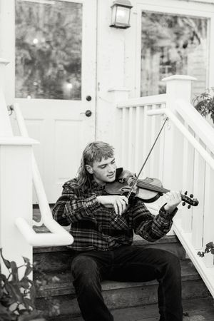 Musician playing violin while sitting on wooden porch steps in black and white photo.