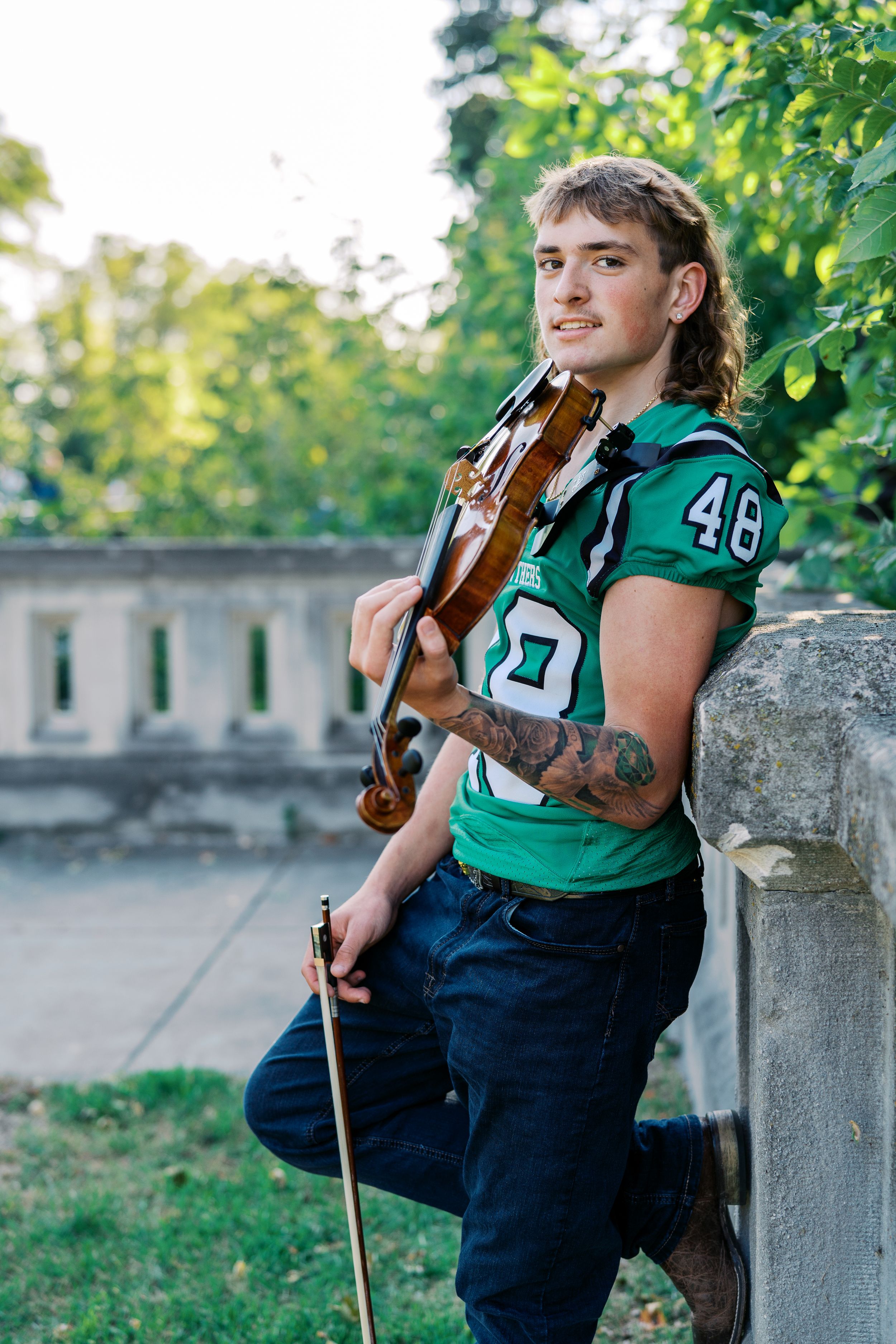 Musician in football jersey playing violin while leaning against stone wall outdoors.