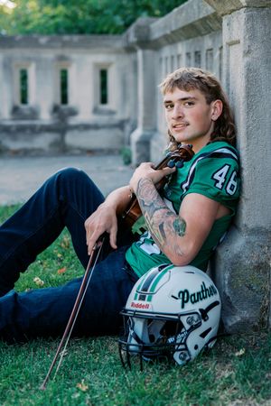 Football player relaxing with violin against stone wall in garden setting.