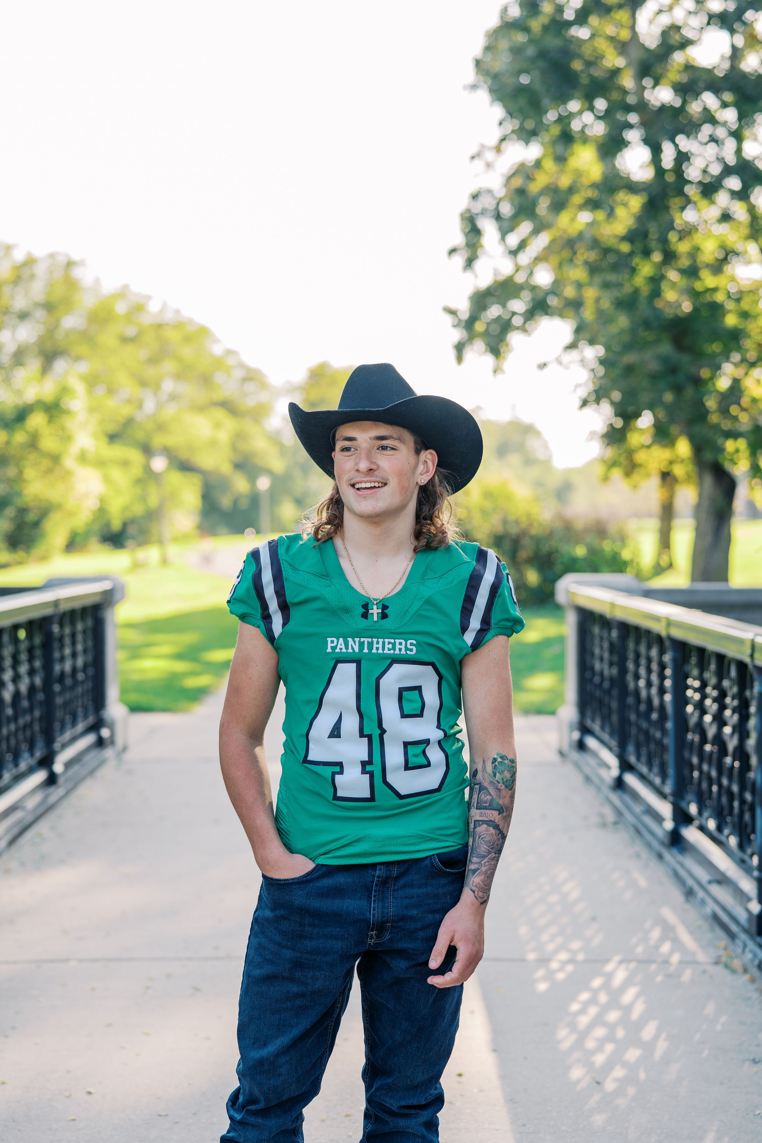 Person in green Panthers football jersey and cowboy hat poses on bridge walkway.