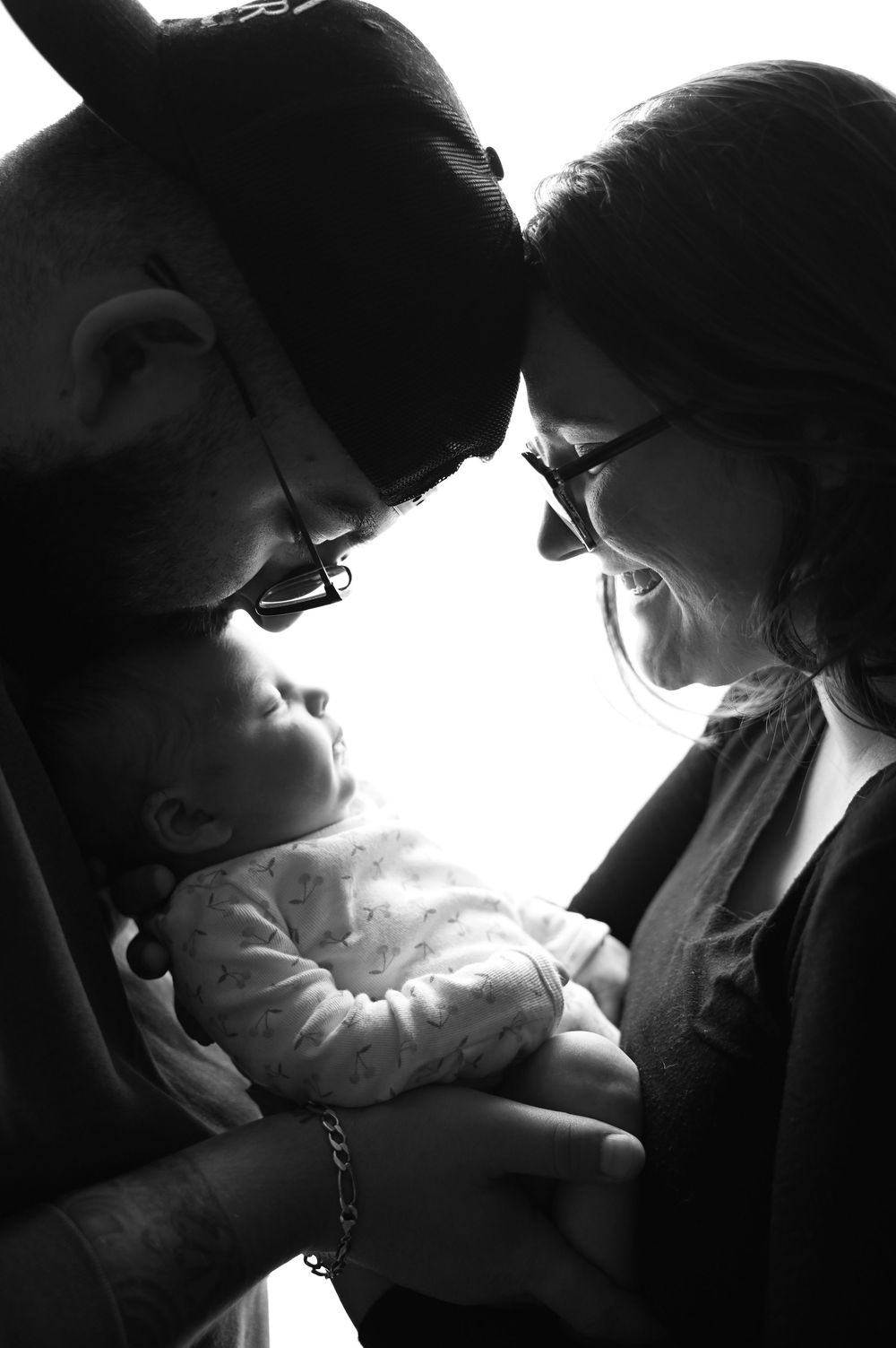 A black and white intimate family portrait showing tender moment between parents and baby in silhouette lighting.