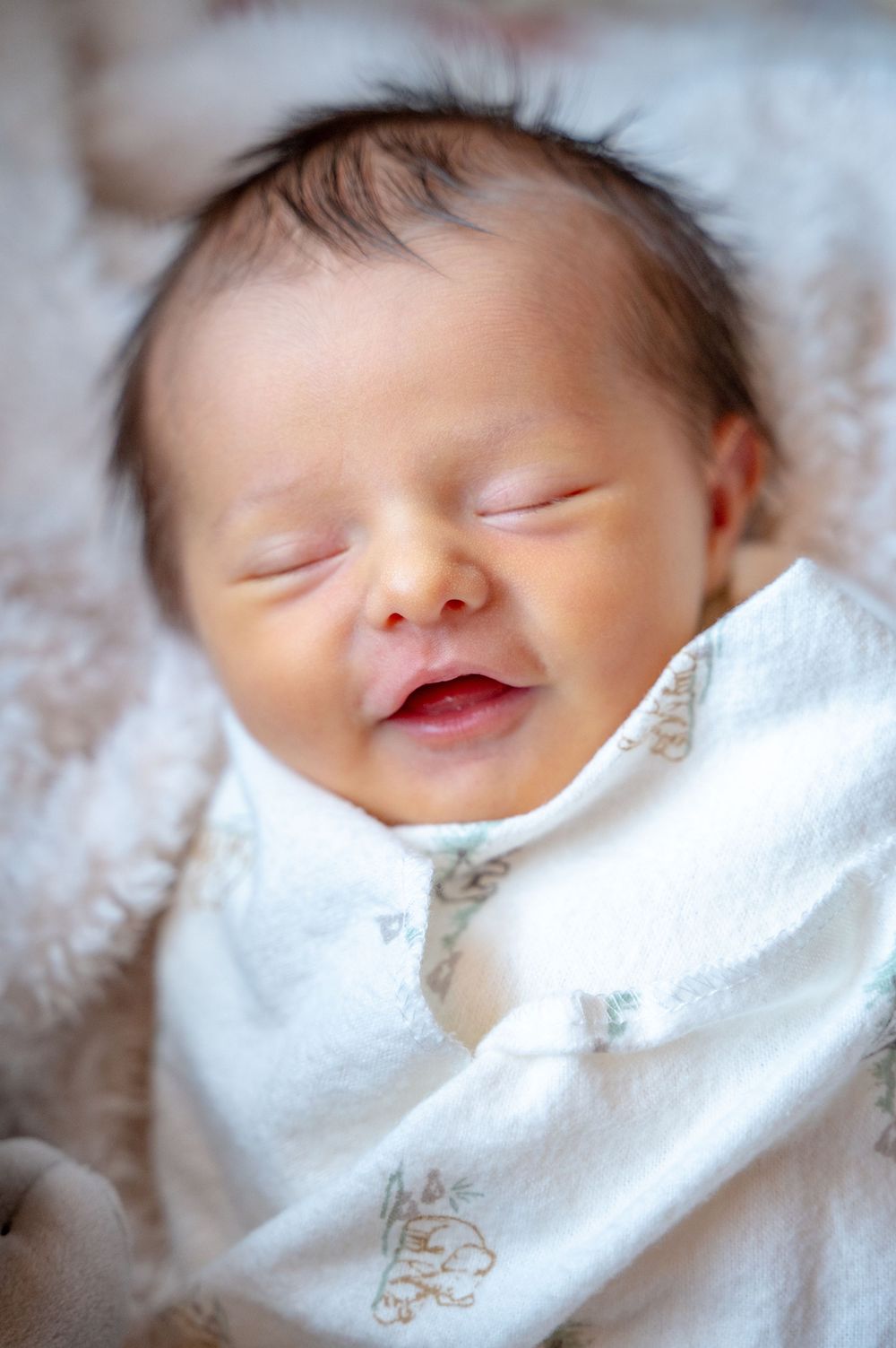 A sleeping newborn baby wrapped in a soft white blanket smiles peacefully while resting.