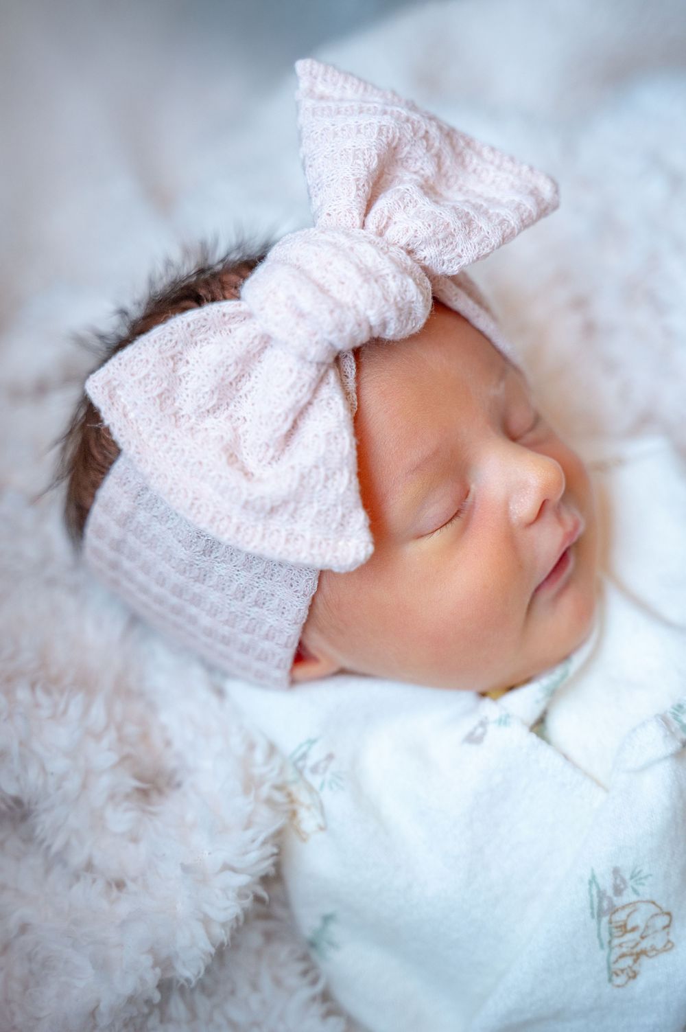 A sleeping newborn wearing a soft pink bow headband rests peacefully on white fluffy bedding.