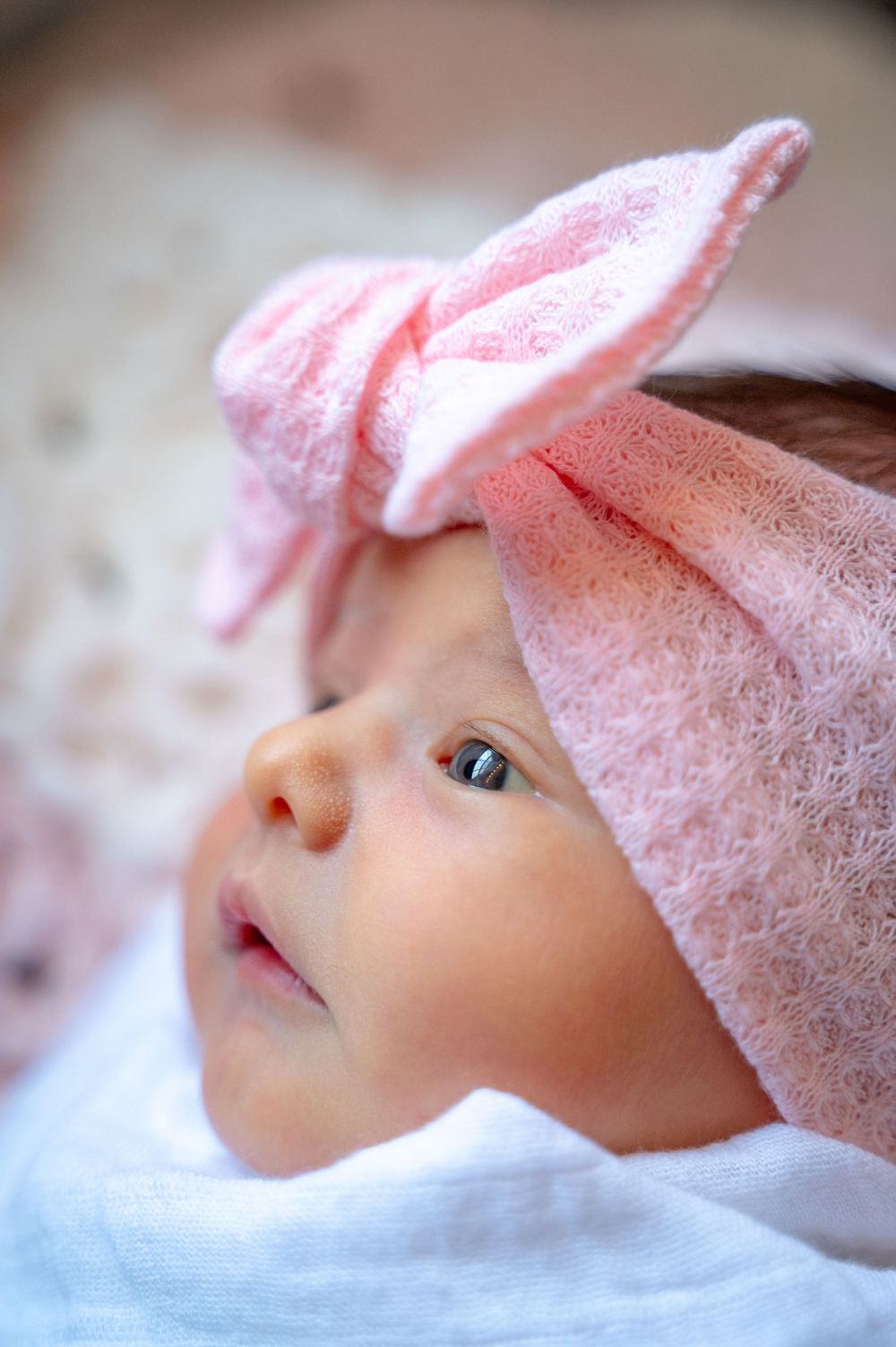 A baby wearing a soft pink lace bonnet gazes upward with bright eyes while resting on white fabric.
