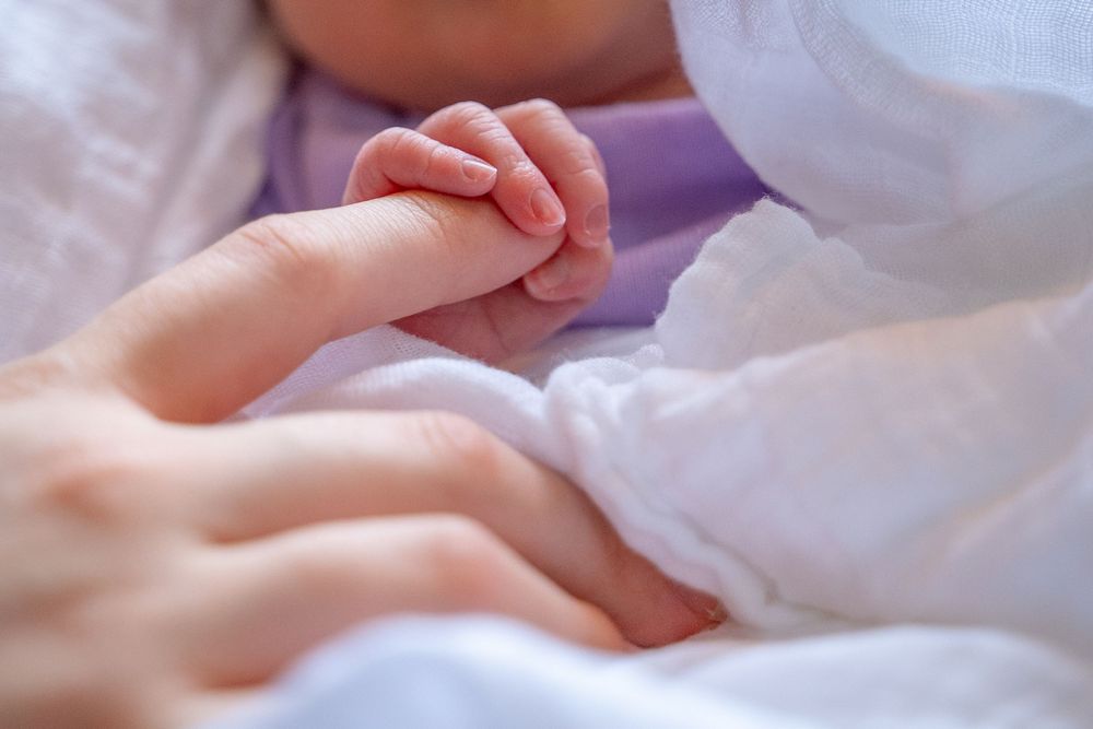 A tiny baby hand grasps an adult finger while resting on soft white bedding.
