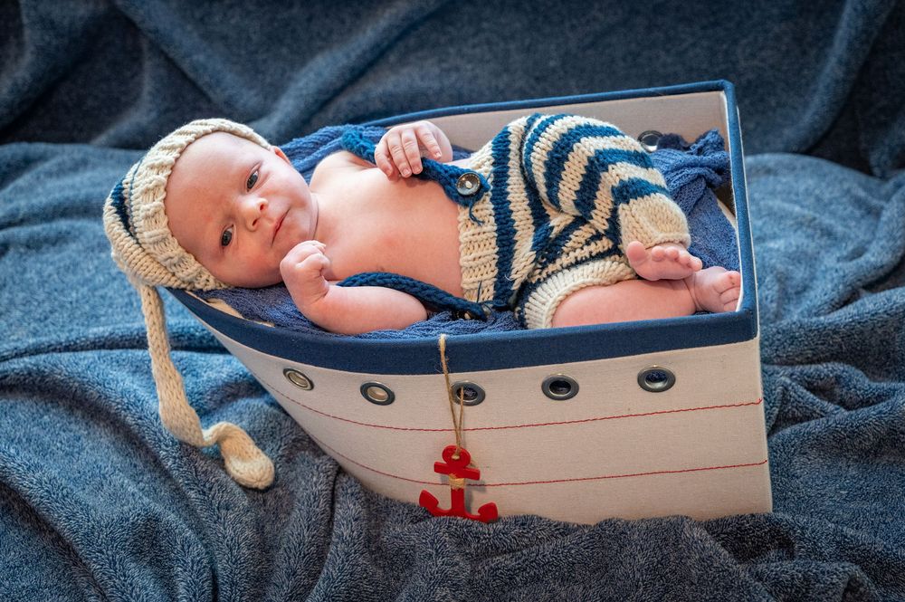 Newborn baby wearing striped nautical outfit lies peacefully in decorative white wooden boat prop against blue backdrop.