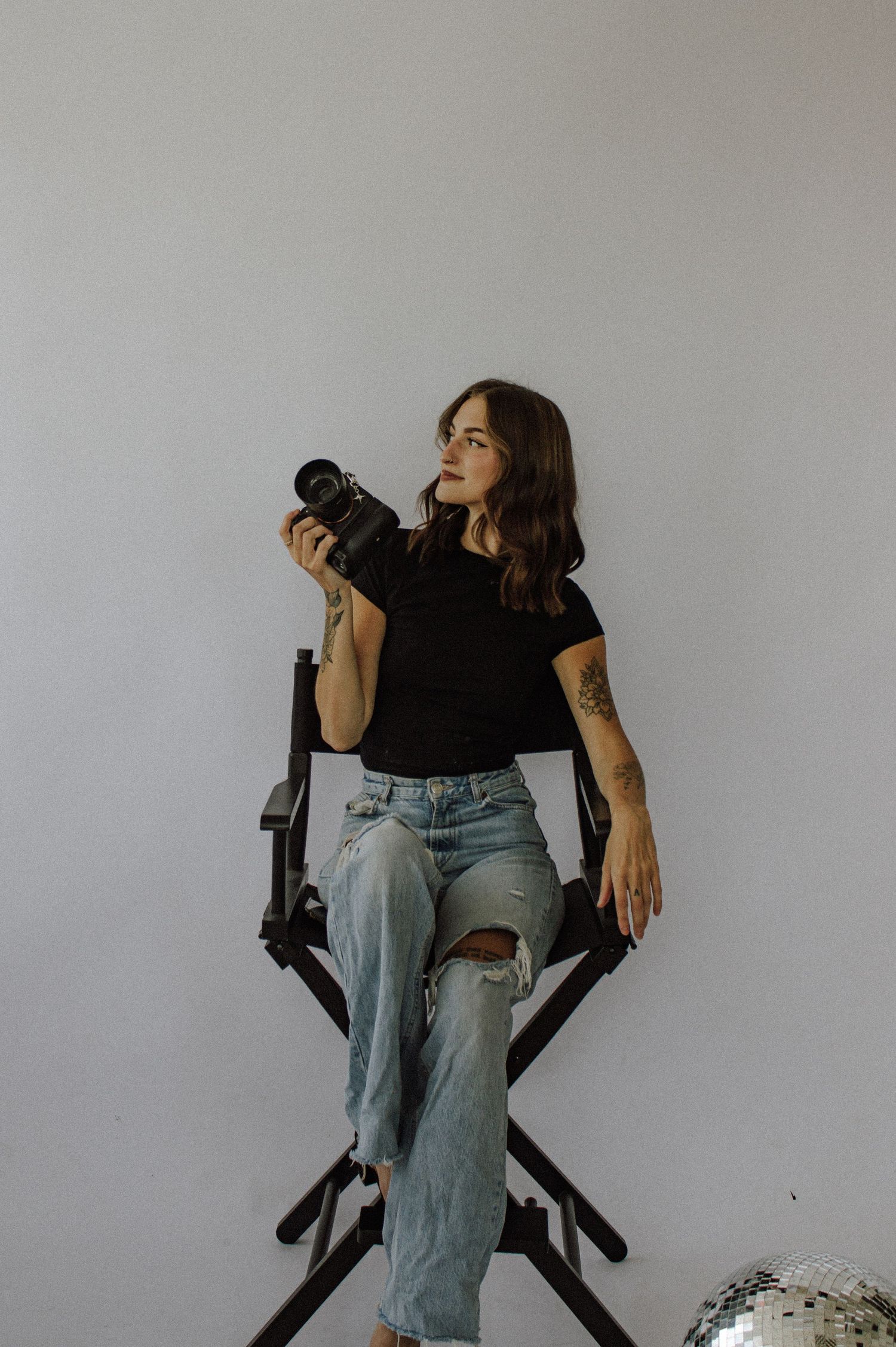 Woman with tattoos in black tee and ripped jeans sits in director's chair holding DSLR camera near disco ball. Kingsport TN