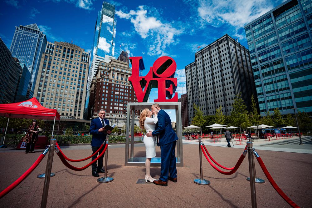 Red LOVE sculpture installation in Philadelphia's Love Park with city buildings and blue sky in the background.
