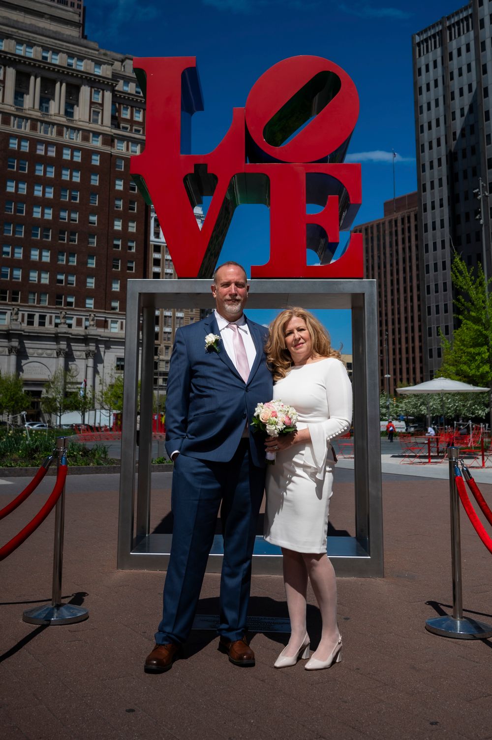 A couple poses for photos in front of the iconic red LOVE sculpture in a city plaza on a sunny day.