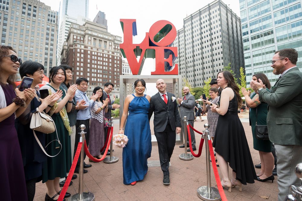 Wedding party gathers under iconic red LOVE sculpture in Philadelphia's Love Park with city buildings in background.