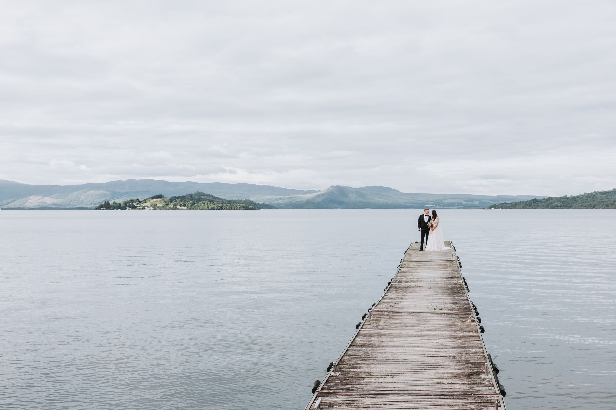A couple stands at the end of a long wooden pier overlooking a misty lake surrounded by mountains.