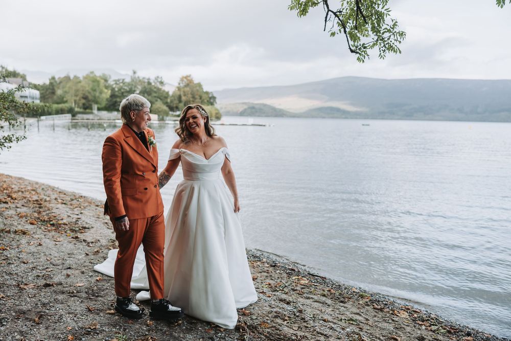 Two people in wedding attire walk along a scenic lakeside beach with mountains in the background.