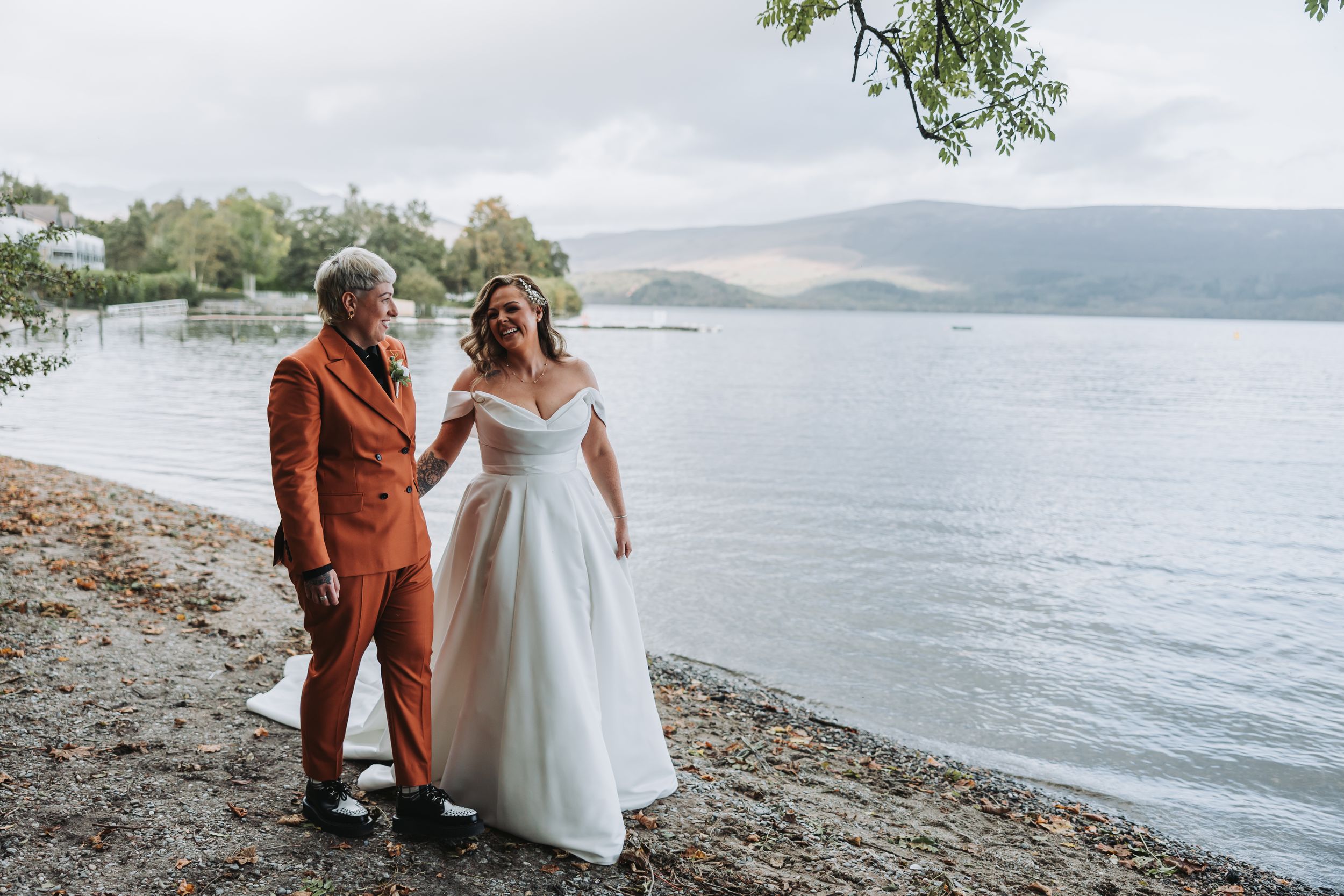 Two people in wedding attire walk along a scenic lakeside beach with mountains in the background.