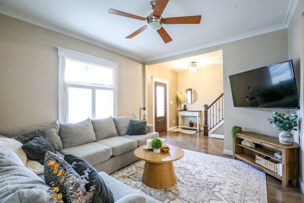 Modern living room with gray sectional sofa, ceiling fan, hardwood floors, and natural light from large windows.