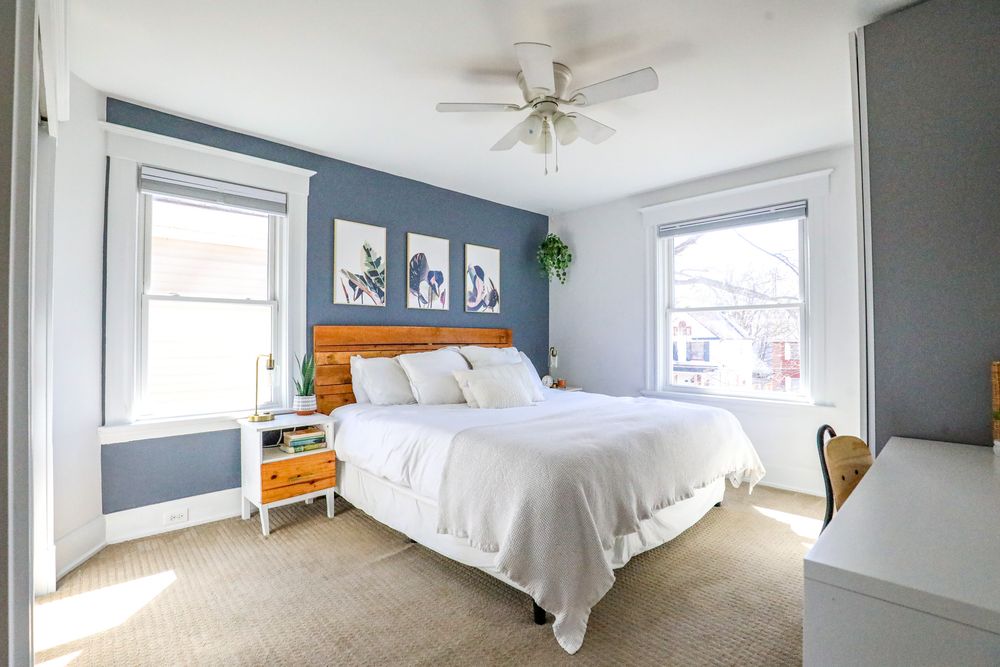 Spacious bedroom with white bedding, ceiling fan, and natural light streaming through multiple windows.