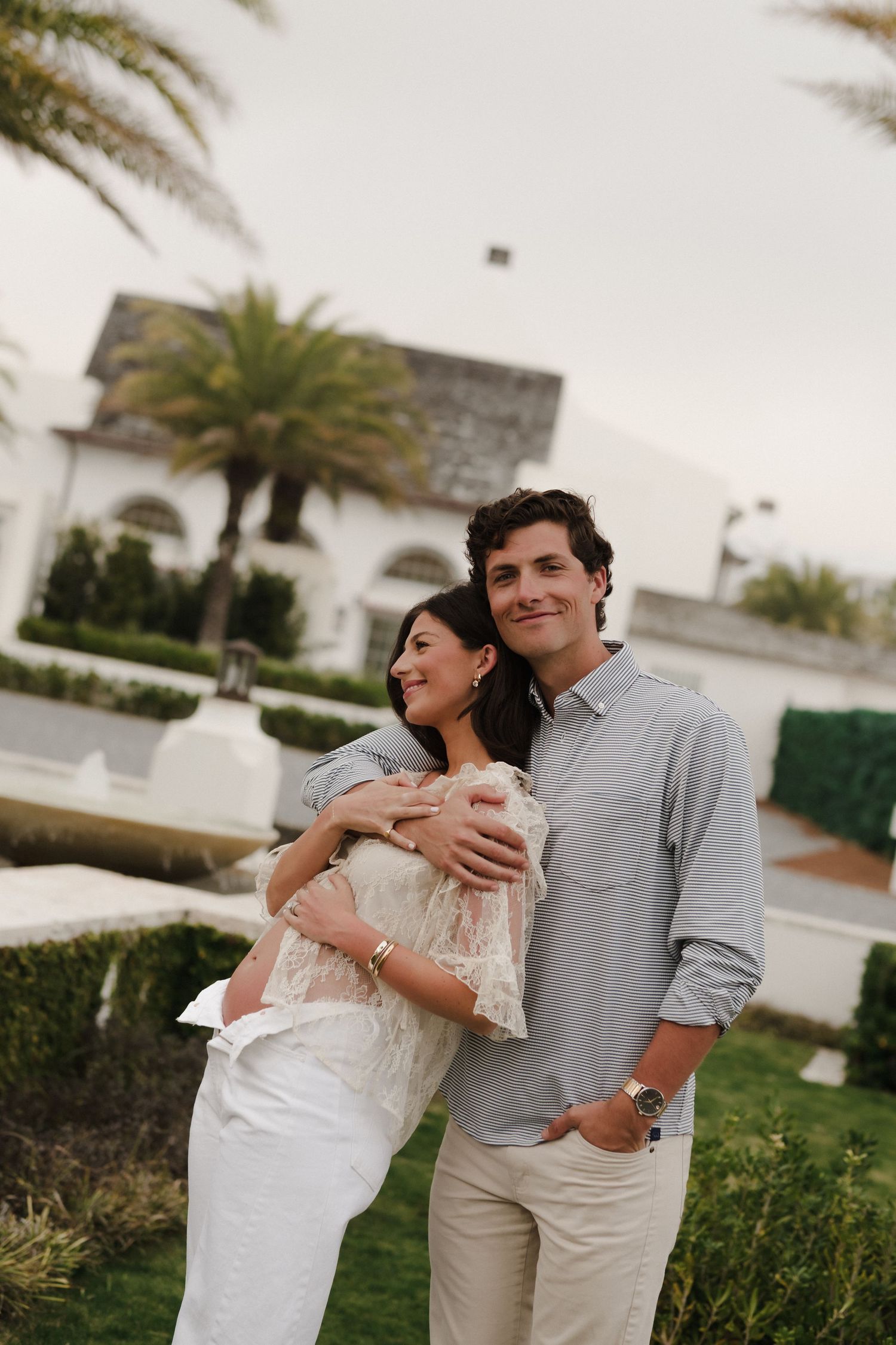 A young couple embracing outdoors near a white building with palm trees, the woman in white and man in a gray shirt.