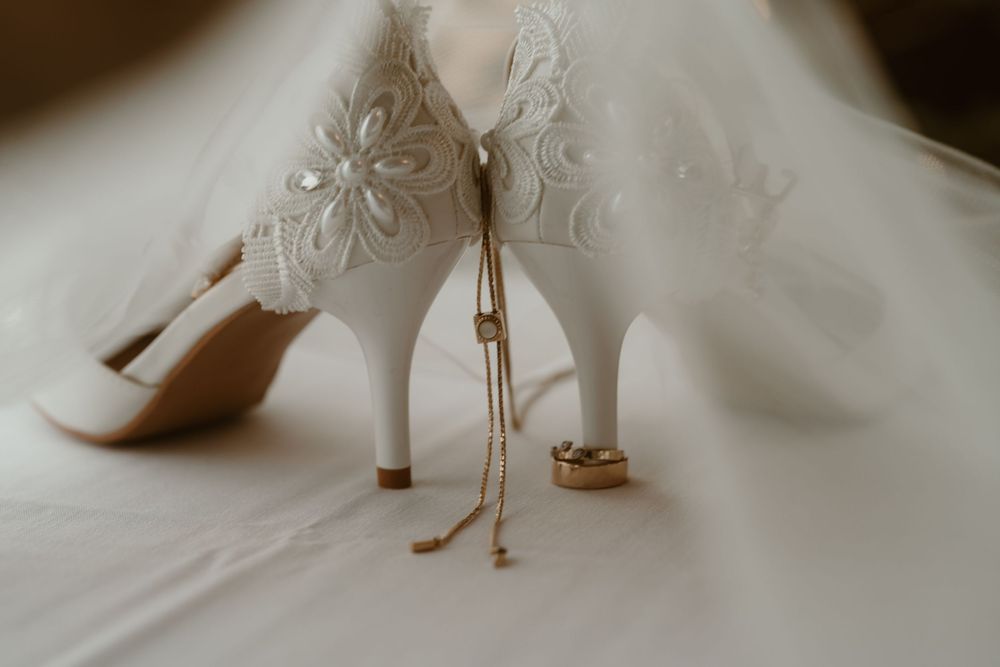 White lace bridal heels with gold jewelry and wedding rings on white fabric.