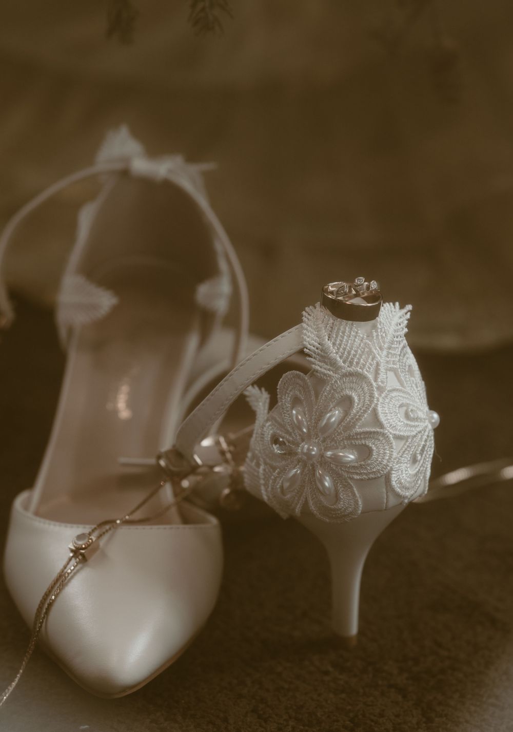 White wedding heels with lace detail and floral embroidery displayed with wedding rings resting on top.