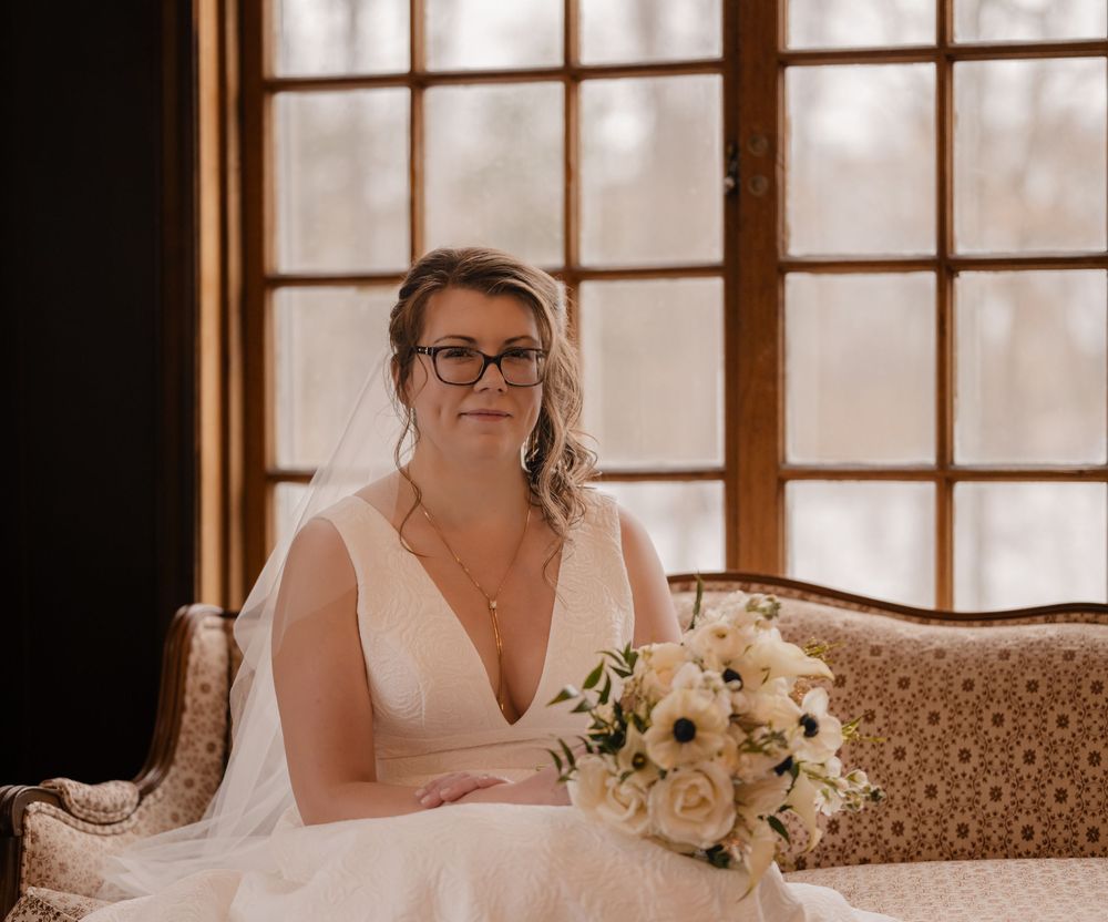 Bride in white V-neck wedding gown holding bouquet of white flowers while seated near large windows.