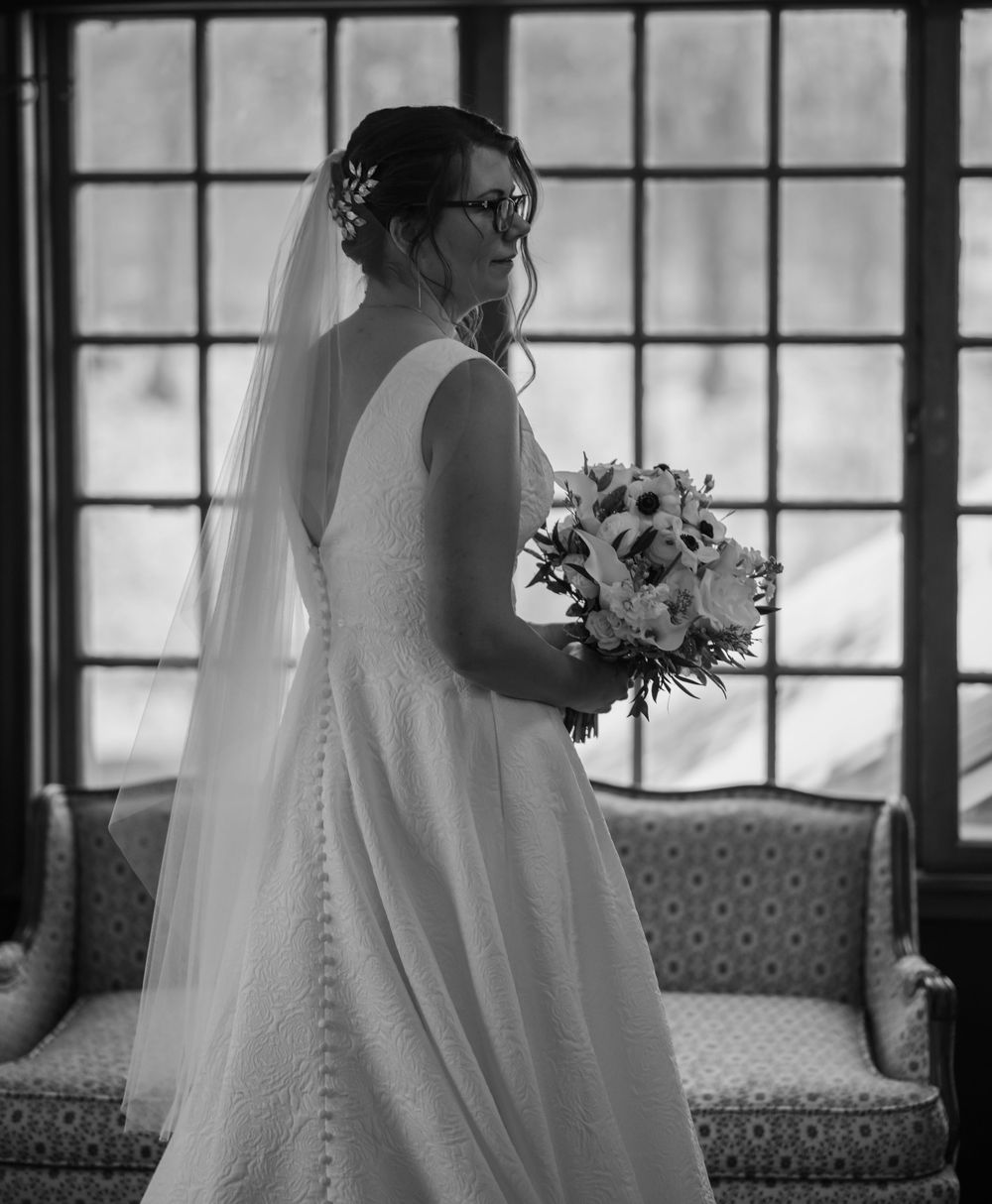 Bride in elegant white gown holding floral bouquet standing by large windows in black and white wedding photo.