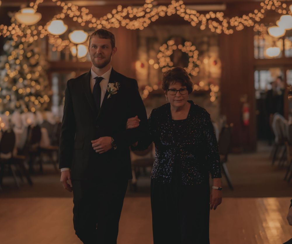 A groom in a black tuxedo escorts an older woman in a navy dress during a wedding reception with warm string lights.