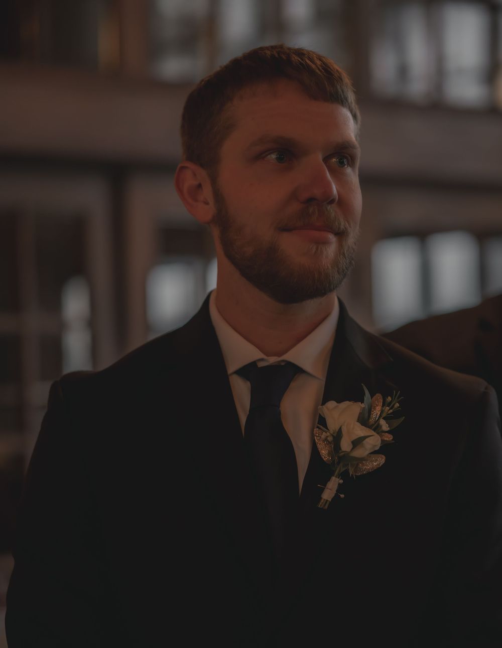 Groom in black suit with floral boutonniere smiling at wedding ceremony.
