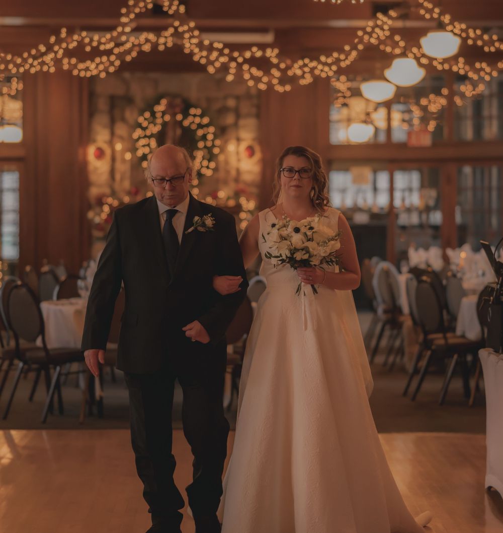 Bride in white gown walks down aisle with older man in dark suit under warm string lights at elegant wedding reception.