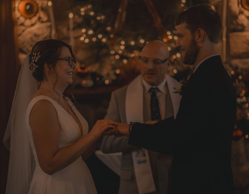 Bride and groom exchange rings during intimate wedding ceremony with warm bokeh lights in background.