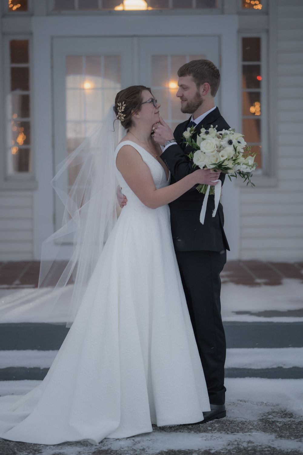 Bride and groom share a romantic moment outdoors in winter, she in a white gown with veil, he holding a bouquet.