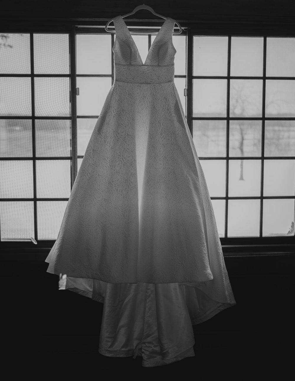 Black and white photo of elegant A-line wedding dress with deep V-neck hanging in window.