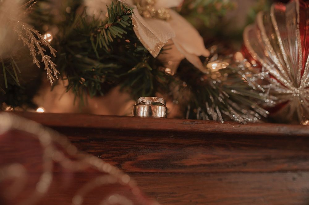 Wedding rings rest on a wooden mantel beneath a decorated Christmas tree with holiday ornaments and greenery.