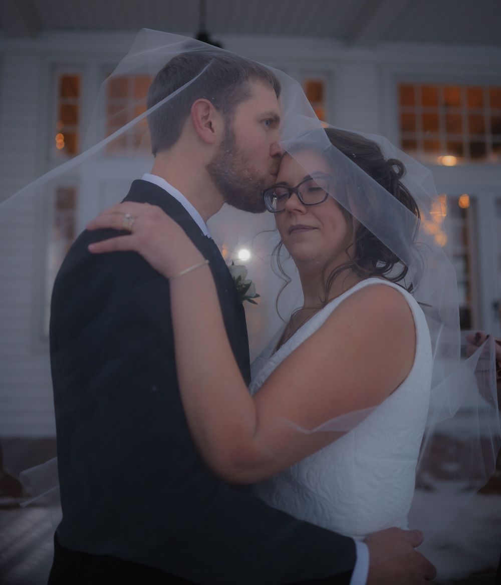 Bride and groom share tender moment under flowing veil, groom kissing brides forehead at wedding reception.