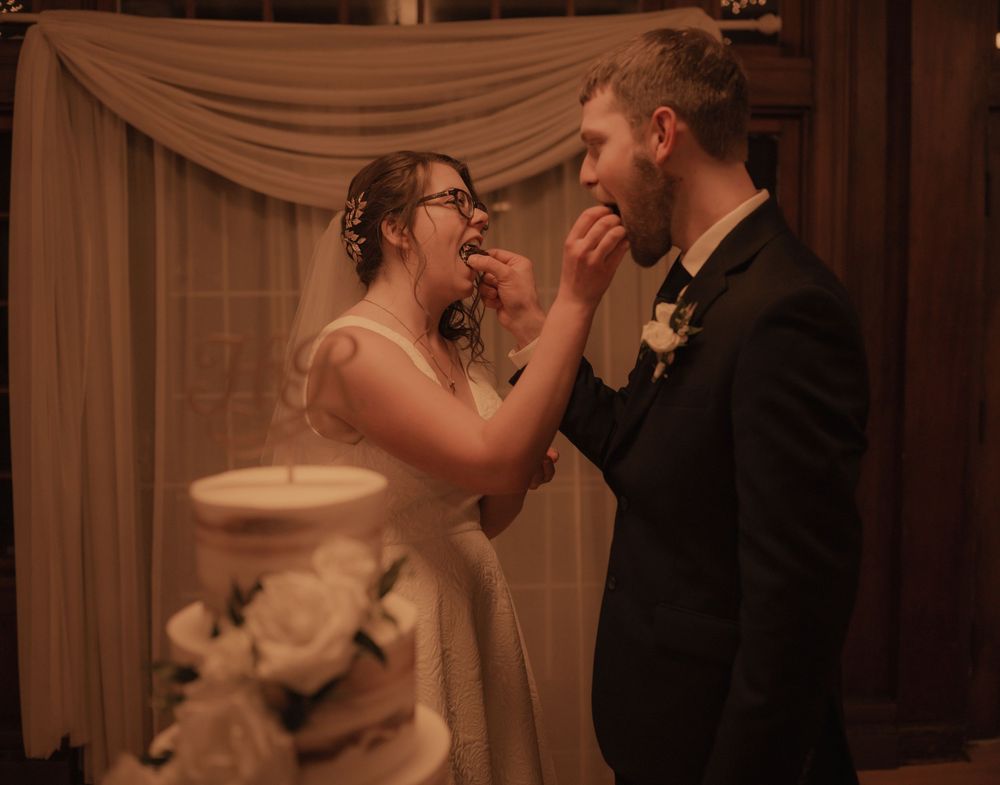 Newlyweds share a joyful cake tasting moment beside elegant white floral wedding cake.