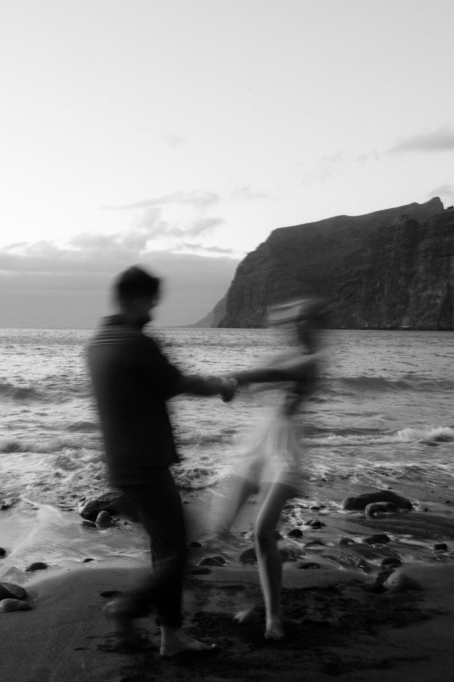 Blurred black and white photo of a couple holding hands on a rocky beach with dramatic cliffs at dusk.