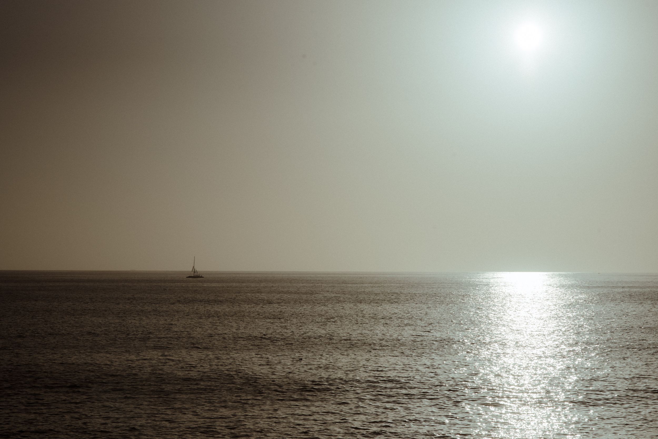 A small sailboat silhouetted against a hazy ocean horizon with sun reflecting off the calm water.