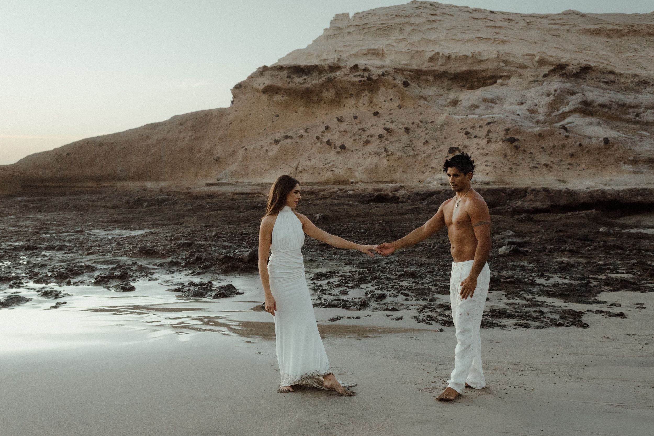 Couple in white outfits walking along a rocky beach coastline with dramatic cliffs in the background at sunset.