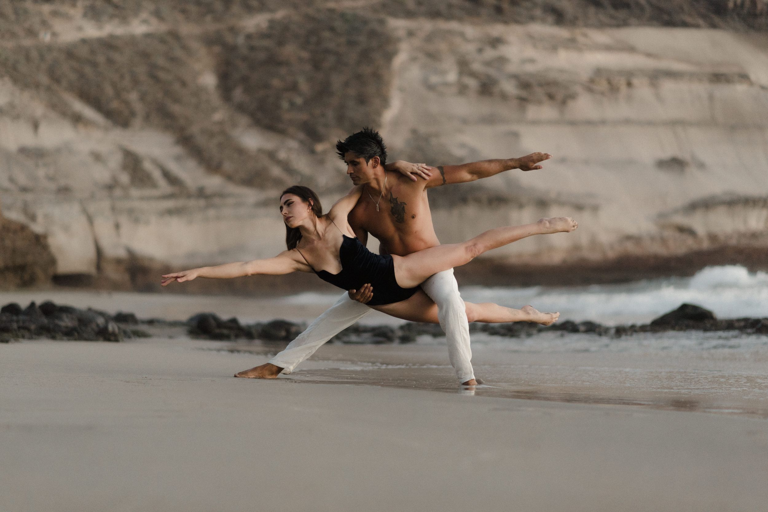 Dancers perform graceful movements on a sandy beach with rocky cliffs and ocean backdrop at dusk.